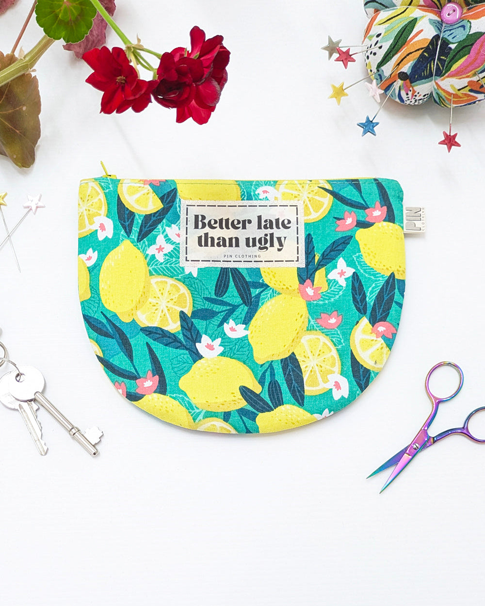 A semicircular fabric pouch with a colorful lemon and leaf print, featuring a white label that reads 'Better late than ugly'. The pouch is styled in a flat lay with red flowers, scissors, keys, and a sewing thread spool on a white background.