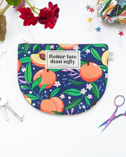 A fabric pouch with a dark blue background and a colorful print of peaches, green leaves, and small white flowers. It features a white rectangular label in the center that reads 'Better late than ugly'.