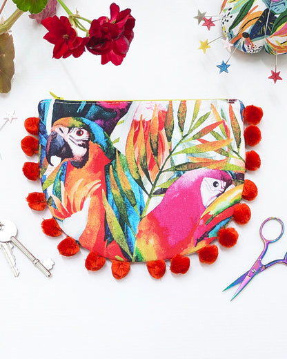 A colorful zippered pouch featuring a vibrant parrot and tropical leaf print, decorated with bright red pom-poms around the border, styled on a white surface with decorative props.